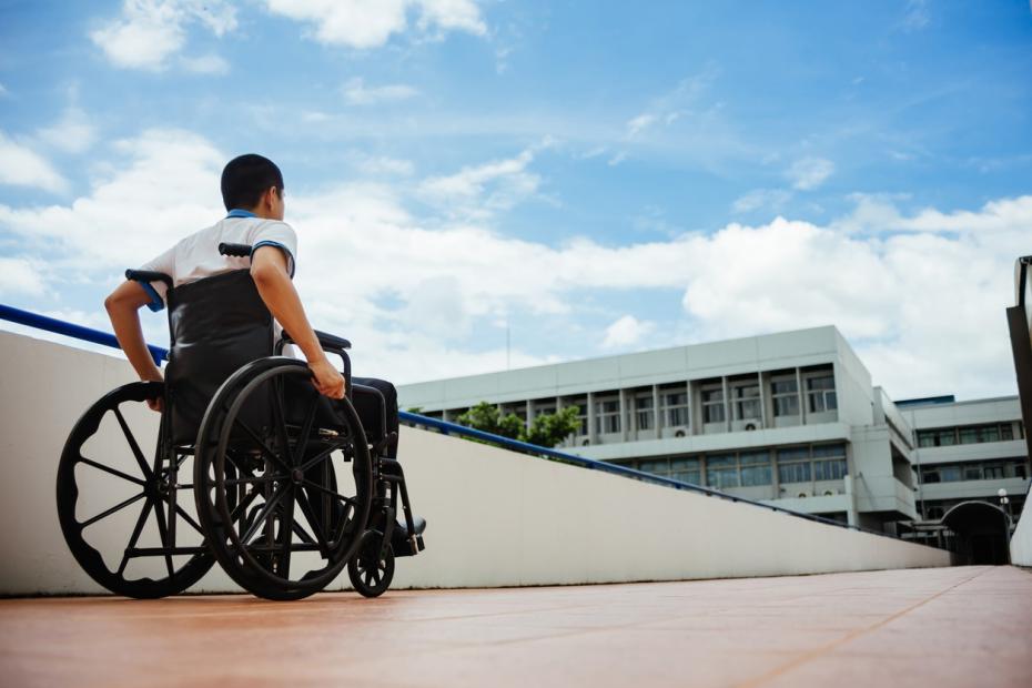 Young man in wheelchair in city wheeling up an incline toward a white office building in the background.