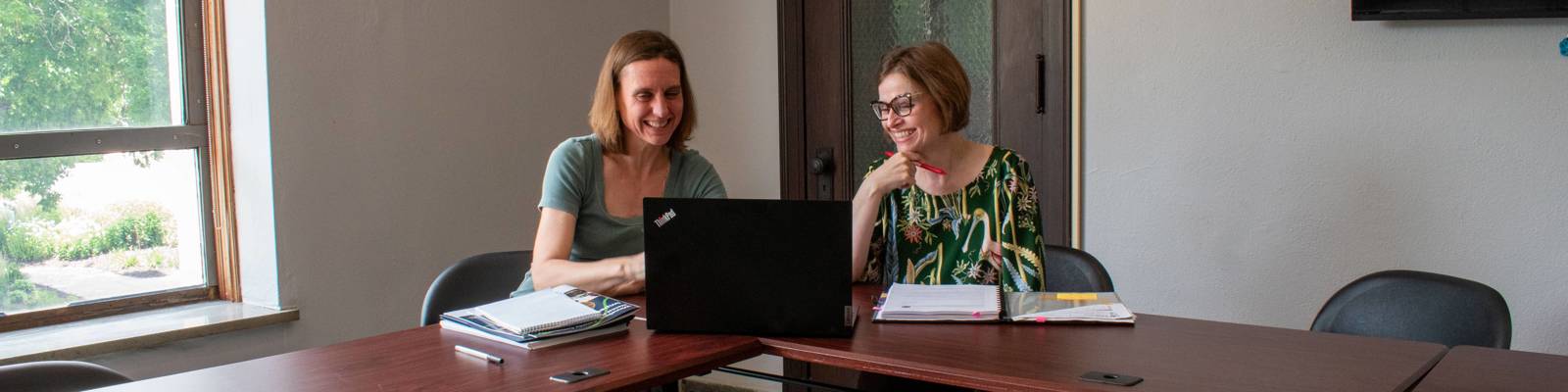 two professional women sitting at conference table in office ooking at laptop with documents piled on table