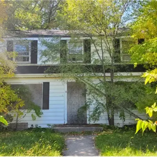 Two-story white house with black shutters and overgrown trees in front yard 