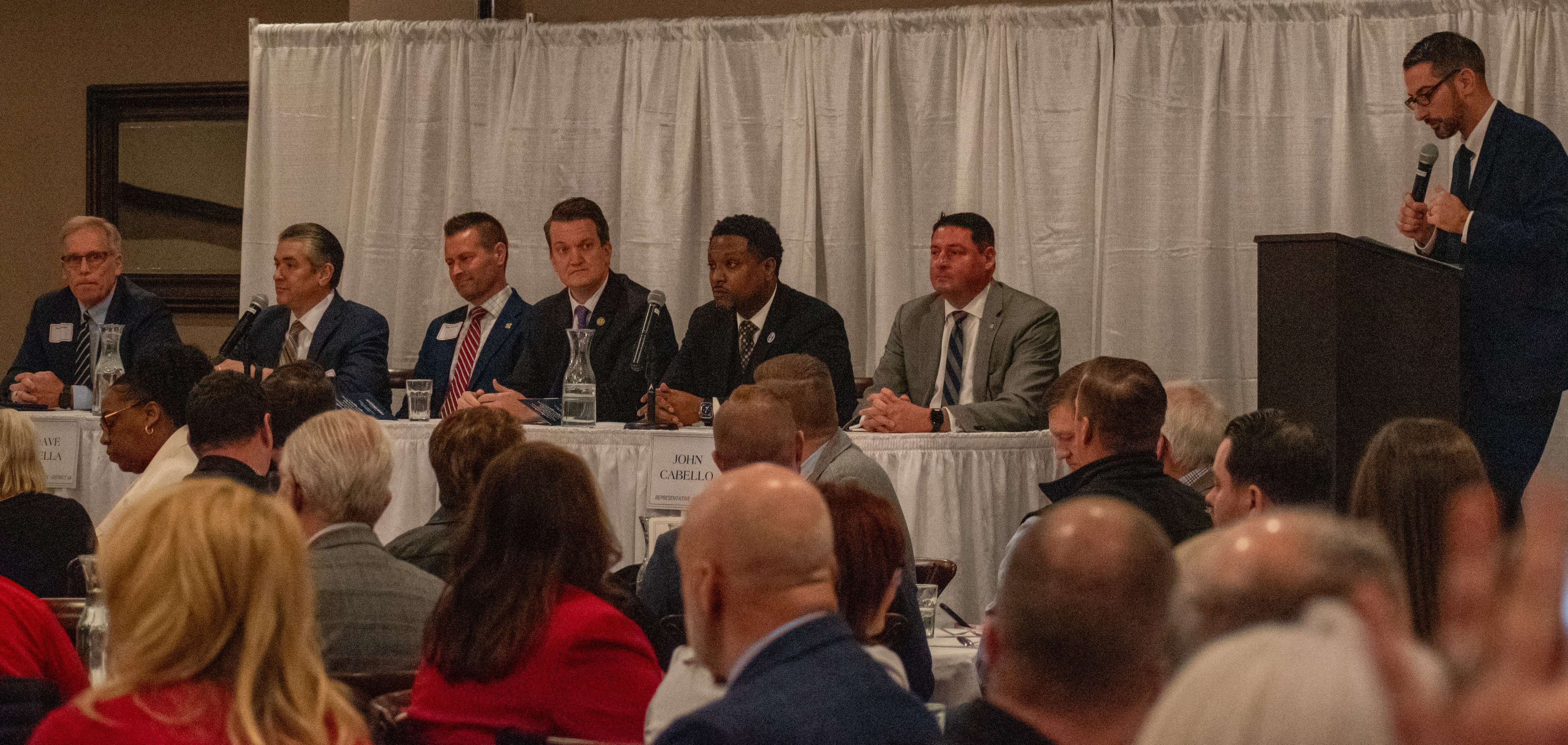 Ballroom set for a luncheon with guests in foreground. Row of legislators seated on stage. From left: Senator Dave Syverson, Rep Dave Vella, Rep Joe Sosnowski, Senator Andrew Chesney, Rep Maurice West II, Rep John Cabello, and facilitator Rockford mayor Tom McNamara at podium.