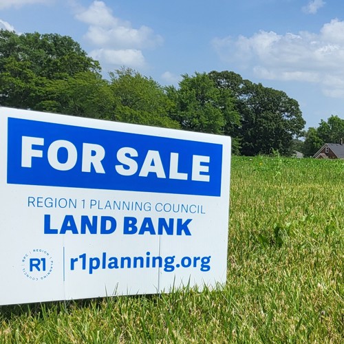 Sign in bright grassy lot that reads For Sale Region 1 Planning Council Land Bank. r1planning.org